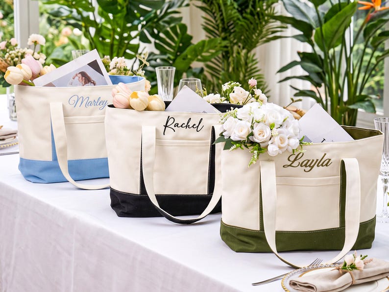 May include: Three canvas tote bags with exterior pockets, each featuring a personalized name embroidered in script. The bags have cream-colored upper sections and contrasting lower panels in light blue, black, and olive green.