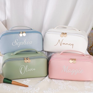 May include: Four personalized cosmetic bags in pastel colors: light blue, white, green, and pink. Each bag has a handle, a gold zipper, and a name embroidered in cursive: Sophia, Nancy, Olivia, and Maggie. The bags are on a white surface.