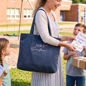 May include: A dark blue corduroy tote bag featuring the embroidered text "MRS. Sinclair" in a light pink script font. The bag has two shoulder straps and a rectangular shape, with a small embroidered butterfly detail next to the name.