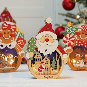 以下が含まれることがあります： 木製のクリスマスオーナメント。ジンジャーブレッドマンとサンタクロースの形。各オーナメントは、内側に祝祭のシーンがあり、小さなライトで照らされています。名前がベースに刻まれています。