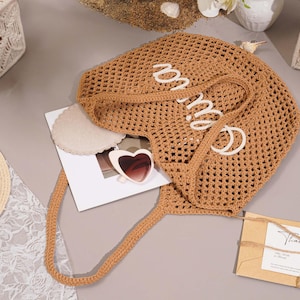 May include: A tan crocheted tote bag with the word "mia" in white script. The bag has long handles and is next to a pair of heart-shaped sunglasses, a straw hat, and a small card that says "Thank You".