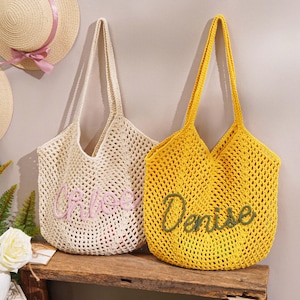 May include: Two crochet tote bags, one cream and one yellow, with long handles. The cream bag has "Chloe" embroidered in pink, and the yellow bag has "Denise" in green. Straw hats with ribbons hang in the background.