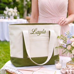 May include: A cream-colored canvas tote bag featuring an olive green base and matching olive green embroidered text that reads Layla. The bag includes two cream-colored shoulder straps and a front exterior pocket, constructed with durable fabric.