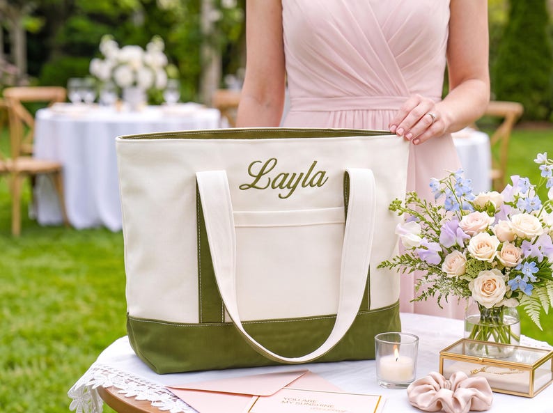 May include: A cream-colored canvas tote bag featuring an olive green base and matching olive green embroidered text that reads Layla. The bag includes two cream-colored shoulder straps and a front exterior pocket, constructed with durable fabric.