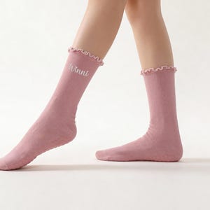 May include: A pair of light pink socks with a ruffled top edge. The word "Winni" is embroidered in white on one sock. The socks are made of a soft material and are knee-high.