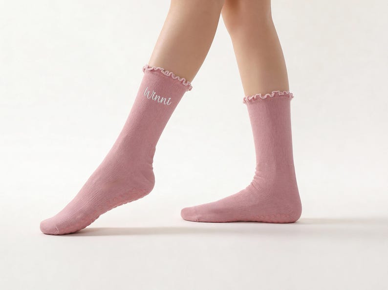 May include: A pair of light pink socks with a ruffled top edge. The word "Winni" is embroidered in white on one sock. The socks are made of a soft material and are knee-high.