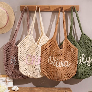 May include: Four crocheted tote bags in shades of mauve, cream, tan, and olive green hang on a wooden rack. Each bag features a different name embroidered in a contrasting color: Hope, Chloe, Olivia, and Lily. Two straw hats with pink ribbons are visible.