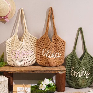 May include: Three open-weave tote bags in cream, brown, and olive green, each with a name embroidered on the front. The bags are displayed on a wooden bench with straw hats hanging in the background. The names are Chloe, Olivia, and Emily.