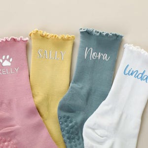 May include: Four pairs of personalized socks in pastel colors: pink, yellow, teal, and white. Each sock features a name embroidered in a complementary color. The pink sock has a paw print design. The socks have a ruffled top and non-slip grips.