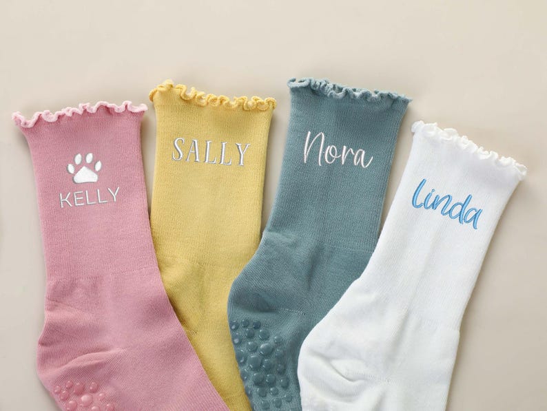 May include: Four pairs of personalized socks in pastel colors: pink, yellow, teal, and white. Each sock features a name embroidered in a complementary color. The pink sock has a paw print design. The socks have a ruffled top and non-slip grips.