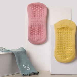 Könnte beinhalten: Drei Paar Socken sind abgebildet. Zwei Paare werden vertikal dargestellt und zeigen die Sohlen mit rutschfesten Griffen. Ein Paar ist rosa, das andere gelb. Ein drittes Paar türkisfarbene Socken mit den Initialen "RS" wird horizontal dargestellt.