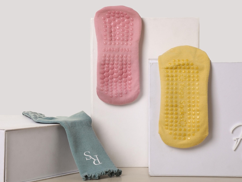 May include: Three pairs of socks in the image. Two pairs are displayed vertically, showing the soles with non-slip grips. One pair is pink, the other is yellow. A third pair of teal socks with the initials "RS" is displayed horizontally.