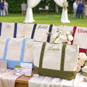 May include: Five canvas tote bags in cream with colored bases and handles in navy, black, burgundy, light blue, and olive green. Each bag features personalized embroidery with names or initials including A, Rachel, Olivia, Marley, and Layla.