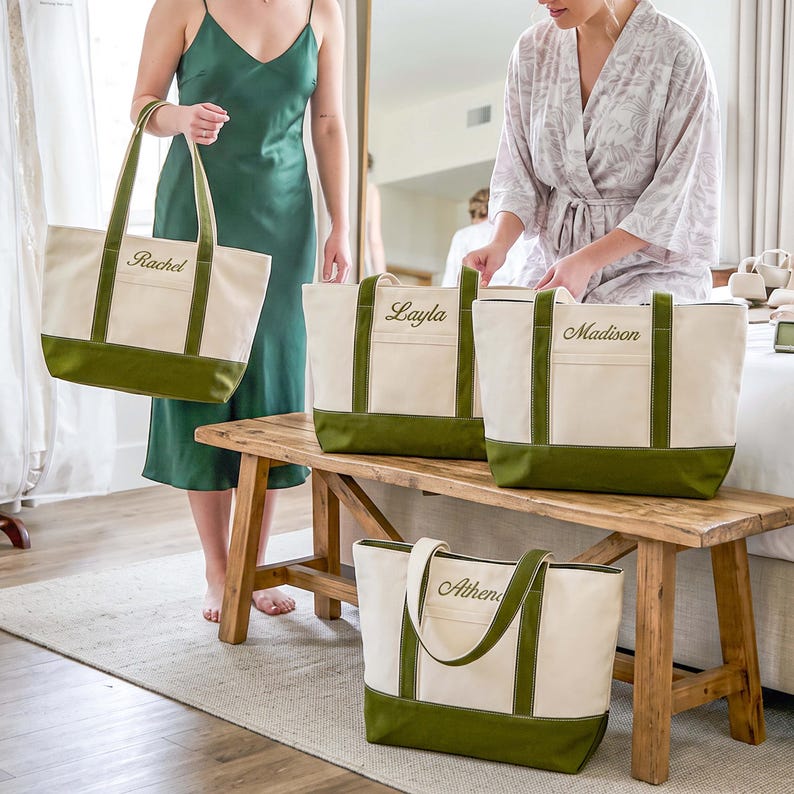 May include: Four cream-colored canvas tote bags with olive green bases and handles. Each bag features a front pocket and is embroidered with a name in cursive script: Rachel, Layla, Madison, and Athena. The bags have a structured rectangular shape.