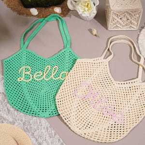 May include: Two crocheted tote bags. One is mint green with "Bella" in cream-colored script. The other is cream-colored with "Chloe" in pink script. Both bags have open weave construction and matching handles. The bags are on a gray surface.