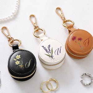 May include: Three round, zippered jewellery cases with gold-tone hardware. Each case features a floral design and a single initial. The cases are black, white, and brown. A pearl necklace, earrings, and rings are also visible.