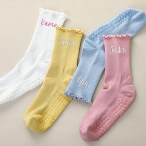 May include: Four pairs of personalized socks in white, yellow, light blue, and pink. Each sock has a ruffled top and a non-slip sole. The socks are embroidered with names: "Kama", "Bride", "Hedy", and "Jodie".