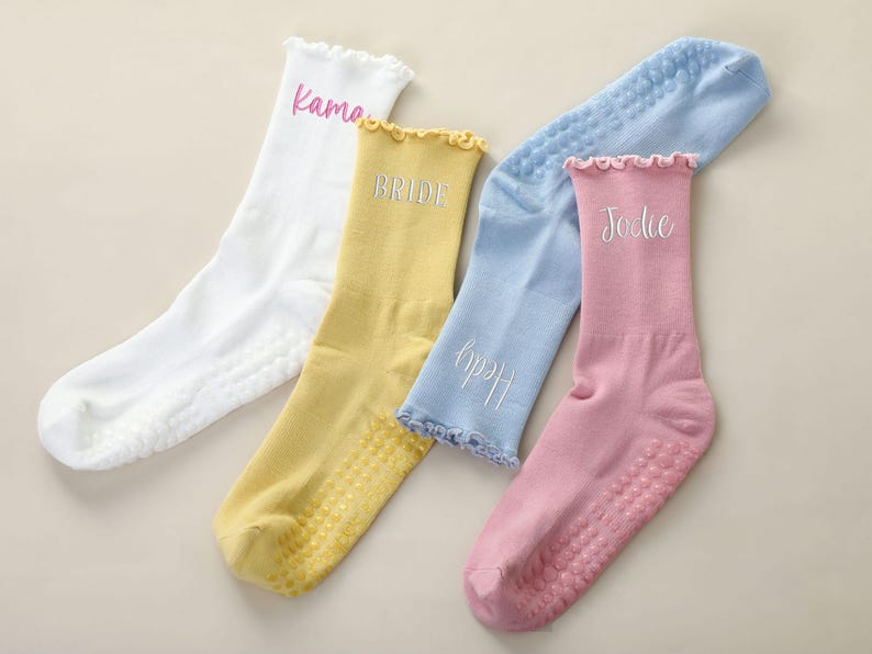 May include: Four pairs of personalized socks in white, yellow, light blue, and pink. Each sock has a ruffled top and a non-slip sole. The socks are embroidered with names: "Kama", "Bride", "Hedy", and "Jodie".