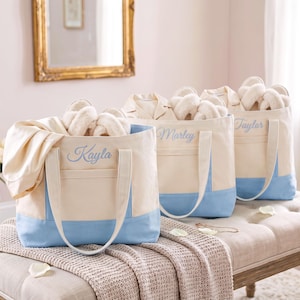 May include: Three cream-colored canvas tote bags with light blue bottom panels and handles. Each bag features a front pocket and is embroidered with a name in light blue script: Kayla, Marley, and Taylor. Each bag contains white slippers.