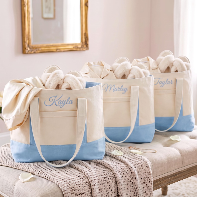 May include: Three cream-colored canvas tote bags with light blue bottom panels and handles. Each bag features a front pocket and is embroidered with a name in light blue script: Kayla, Marley, and Taylor. Each bag contains white slippers.