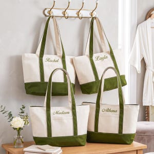 May include: Four off-white canvas tote bags with olive green trim and handles. Each bag features a personalized name embroidered in green script: Rachel, Layla, Madison, and Athena. The bags are displayed hanging on a wall hook and resting on a table.