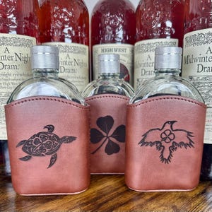 May include: Clear glass flasks with silver caps, encased in brown leather sleeves. Each sleeve features a unique etched design: a turtle, a four-leaf clover, and a bird. Bottles of whiskey are in the background.