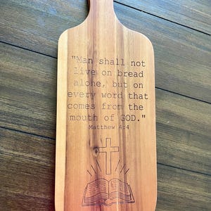 Könnte beinhalten: Holzschneidebrett mit Griff und graviertem Text. Der Text lautet: „Der Mensch lebt nicht vom Brot allein, sondern von jedem Wort, das aus dem Mund Gottes kommt.“ Matthäus 4,4. Ein Kreuz und ein offenes Buch sind ebenfalls graviert.