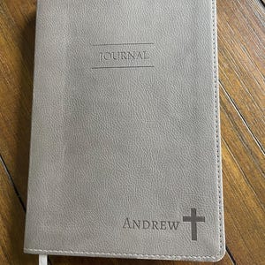 Könnte beinhalten: Ein graues Notizbuch mit dem Wort "JOURNAL" oben eingeprägt. Der Name "ANDREW" und ein Kreuz sind unten rechts aufgedruckt. Ein weißes Lesezeichenband ist sichtbar.
