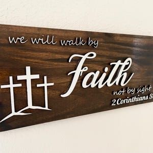 Könnte beinhalten: Dunkel gebeiztes Holzschild mit den Worten "we will walk by Faith not by sight" und dem Bibelvers 2. Korinther 5,7. Der Text und drei Kreuze sind weiß auf dem Holzgrund dargestellt.