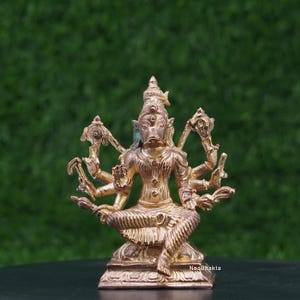 May include: A bronze-colored statue of a multi-armed deity with a boar-like face, seated in a meditative pose. The statue has intricate details, including ornate headwear and jewelry. The background is a blurred green, with the NeoBhakta logo visible.