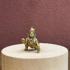 Könnte beinhalten: Eine kleine, goldfarbene Metallfigur, die eine krabbelnde Gottheit darstellt. Die Figur zeigt detaillierte Gesichtszüge, einen kronenartigen Kopfschmuck sowie verzierten Schmuck an Hals und Armen in einer knienden, krabbelnden Haltung.