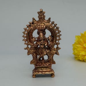 May include: A copper-toned metal figurine depicting a deity playing a flute, positioned within an ornate, circular, flame-like halo frame. The figure stands on a rectangular base, featuring intricate relief work and a polished metallic finish.