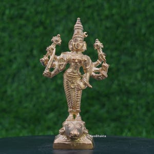 Könnte beinhalten: Eine goldfarbene Statue einer mehrarmigen Gottheit, die auf einem Sockel mit einer Tierfigur steht. Die Statue weist komplizierte Details auf und ist vor einem grünen Hintergrund platziert. Der Text "NeoBhakta" ist auf dem Sockel zu sehen.