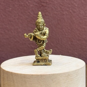 May include: A small, metallic gold-coloured figurine depicting a figure standing on one leg while playing a flute. The figure wears a pointed headdress and is positioned on a small, rectangular base, showcasing intricate details in the metal casting.