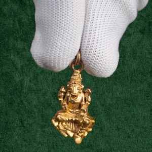 May include: A gold-colored pendant featuring a seated deity with ornate headdress and multiple arms. The pendant is suspended from a small loop, and the figure is detailed with intricate carvings. The background is a dark green fabric.