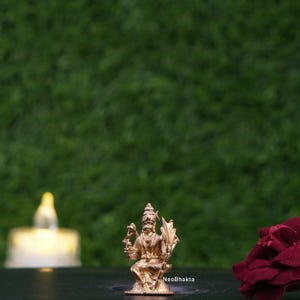 Puede incluir: Pequeña estatua dorada de una deidad sentada con detalles intrincados. La estatua está sobre una superficie oscura, con un fondo verde borroso. El texto "NeoBhakta" es visible en la base. Una vela encendida y una flor roja también están en el fondo.