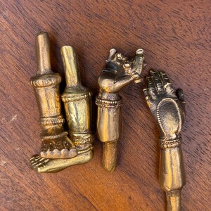 May include: Four separate brass components depicting anatomical parts, including two legs with detailed feet and anklets, and two arms with detailed hands and bracelets. Each piece features a cylindrical extension at the top or bottom end.