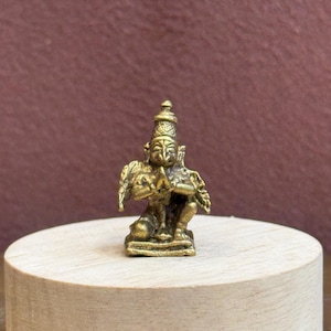 Può includere: Una piccola statuetta metallica color oro raffigurante una divinità inginocchiata con ali, ritratta con le mani giunte in posizione di preghiera. La figura presenta incisioni complesse sul copricapo e sulle ali, poggiando su una piccola base rettangolare.