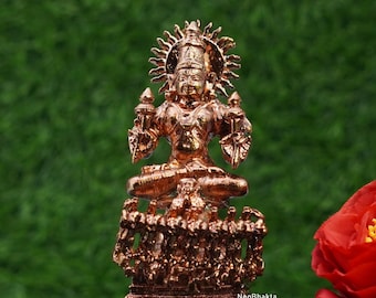 May include: A bronze-colored statue of a seated deity with a halo, intricate details, and multiple arms. The statue is on a base with figures and the text "NeoBhakta". The background is a blurred green.