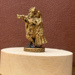 Könnte beinhalten: Eine kleine, goldfarbene Metallfigur, die zwei zusammenstehende Personen darstellt. Eine Figur hält ein flötenartiges Instrument, während die andere eng daneben steht. Die Skulptur weist detaillierte Verzierungen an der Kleidung und Kopfbedeckung auf.