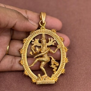 May include: A gold-toned metal pendant featuring a central figure of a multi-armed deity in a dancing pose, enclosed within an oval frame decorated with radiating spikes. The pendant includes a top bail for attachment to a chain or cord.