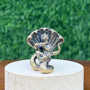 May include: A small, metallic brass-coloured figurine depicting a figure playing a flute while seated atop a coiled multi-headed serpent. The sculpture features intricate detailing on the deity's form and the textured scales of the snake.