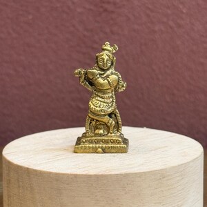 Könnte beinhalten: Eine kleine, goldfarbene Messingfigur, die eine Gottheit in einer überkreuzten Beinhaltung beim Spielen einer Flöte darstellt. Die Figur weist detaillierte Gravuren an Kleidung und Schmuck auf und steht auf einem rechteckigen Sockel.