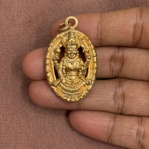 Può includere: Un ciondolo ovale color oro con un rilievo dettagliato di una divinità a più braccia. Il ciondolo presenta un bordo testurizzato e un piccolo anello circolare sulla parte superiore per l'aggancio. La superficie metallica ha una finitura opaca e invecchiata.