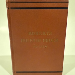 May include: A brown book titled "Roberts Rules of Order Revised" with gold lettering. The book has black horizontal lines near the top and bottom edges. The book is standing upright.