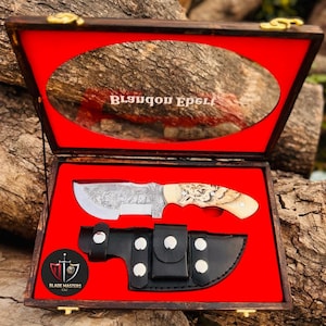 Puede incluir: Un cuchillo personalizado con una hoja decorativa y un mango de hueso, presentado en una caja de madera con interior de terciopelo rojo. Incluye una funda de cuero negro. La caja tiene el texto "Blsuqon Epel" y el logotipo "Blade Masters LLC".