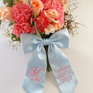 Könnte beinhalten: Ein Blumenstrauß aus rosa und pfirsichfarbenen Rosen und Nelken, akzentuiert mit Schleierkraut. Die Blumen sind mit einem hellblauen Satinband gebunden, das mit dem Buchstaben "M" und den Worten "Ascension HOCO 2025" in Rosa bestickt ist.