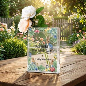 Puede incluir: Un jarrón rectangular transparente con ilustraciones florales y de Peter Rabbit, que contiene rosas blancas y melocotón. El jarrón presenta el texto "Peter Rabbit's Garden" y "A Tale by Beatrix Potter". La escena está ambientada al aire libre.