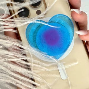 May include: A clear phone case with a gold trim, featuring a blue heart-shaped phone grip. The grip has a gradient effect, transitioning from a deep purple center to a sparkling blue. White feathers are in the background.