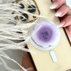May include: A light gold phone with a purple and white phone grip. The grip has a circular design with a purple center and small crystals. White feathers are in the background.